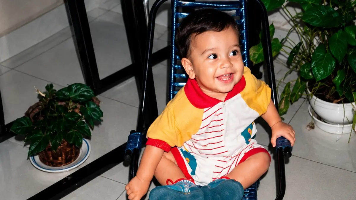 Murilo bebê, sentado numa cadeirinha de fios azuis, sorrindo, de roupinha amarela e vermelha