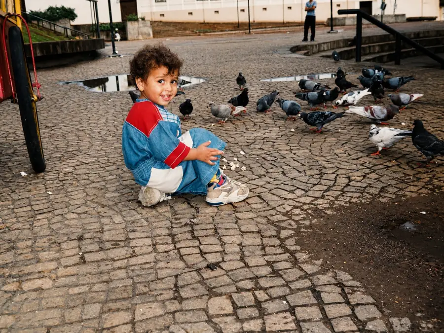 Murilo pequeno, sentado numa praça de pedras portuguesas, sorrindo, com pombos em volta